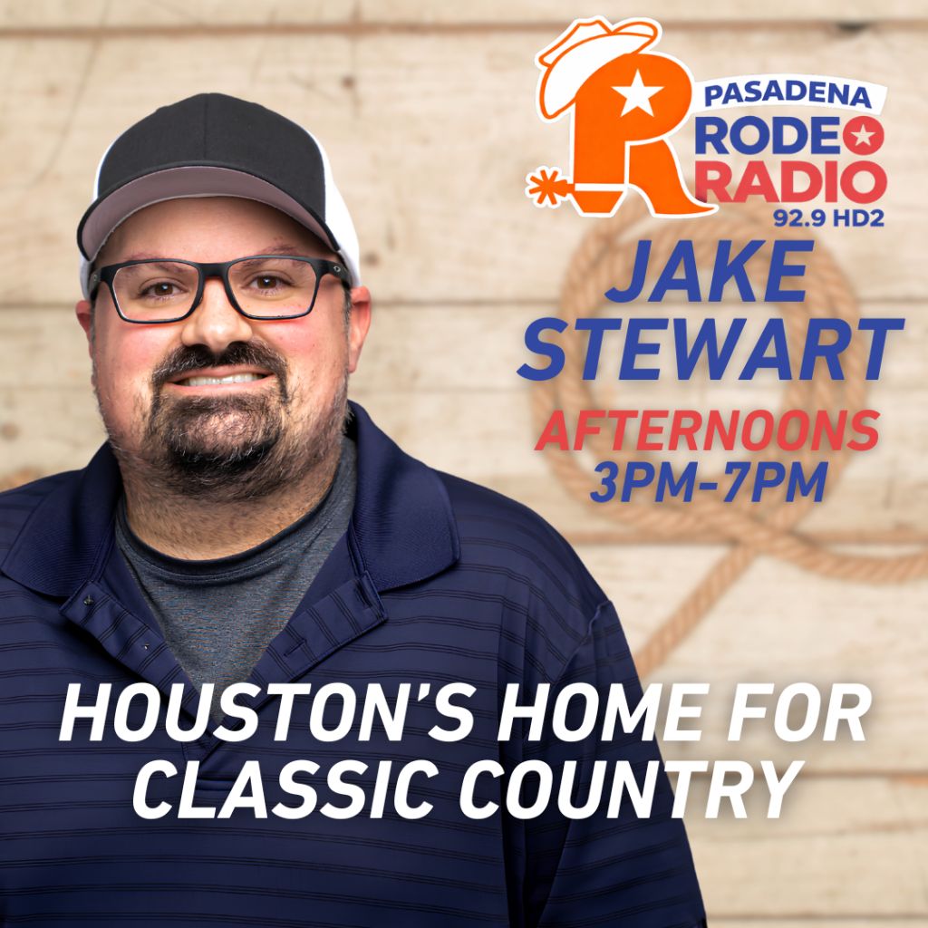 A smiling man with a beard wearing glasses and a baseball cap, standing in front of a wall with the Pasadena Rodeo Radio 92.9 HD2 logo and the text "JAKE STEWART AFTERNOONS 3PM-7PM" and "HOUSTON'S HOME FOR CLASSIC COUNTRY".